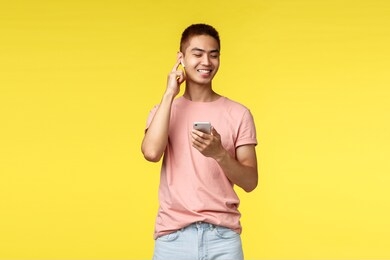 technology, communication and lifestyle concept. handsome asian man using mobile phone and wireless earphones, enjoying nice sound of music, close eyes and smiling, hold smartphone