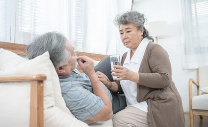 asian senior woman look after give glass of water to elder man taking medicines and drinking water while lie on couch at home, health and medicine concept with copy space.