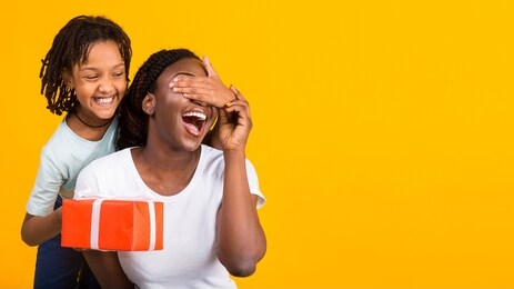 mother's day concept. smiling black child keeping her mom's eyes covered and holding present box, yellow wall, copyspace