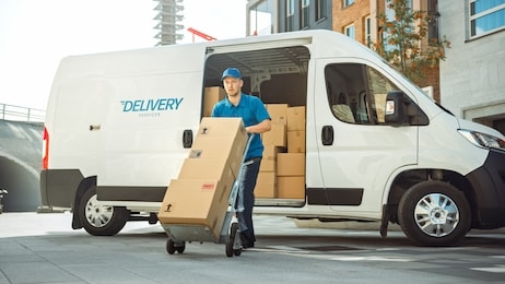 delivery man pushes hand truck trolley full of cardboard boxes hands package to a customer. courier delivers parcel to man in business district.