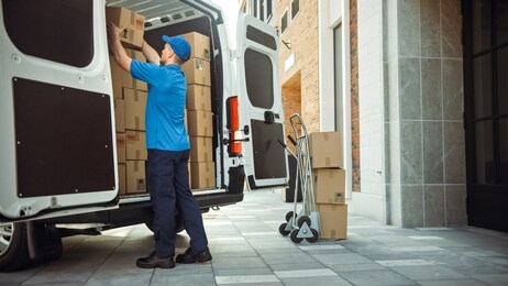 delivery man uses hand truck trolley full of cardboard boxes and packages, loads parcels into truck / van. professional courier / loader helping you move, delivering your purchased items efficiently
