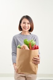 beautiful smiling asian woman holding paper shopping bag full of vegetables and groceries, studio shot isolated on white background