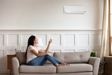 vietnamese young woman relaxing on comfy couch in contemporary living room with air conditioner, holding remote control turning on or off cooler system, setting comfortable temperature at modern home