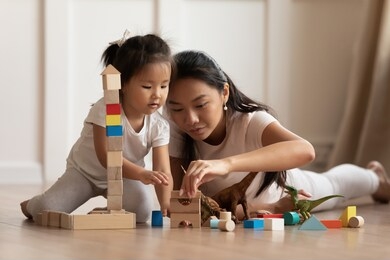 involved little daughter her vietnamese mother play on warm floor using wooden colorful blocks create towers and buildings improve fine motor skill of kid. funny educational games for children concept