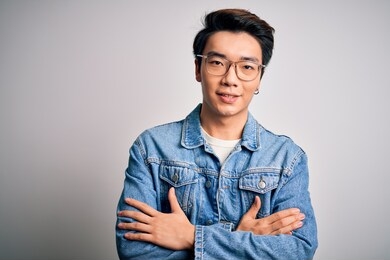 young handsome chinese man wearing denim jacket and glasses over white background happy face smiling with crossed arms looking at the camera. positive person.