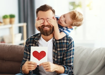 happy young bearded man in casual wear with eyes closed and greeting card while sitting with little son on sofa at home in fathers day
