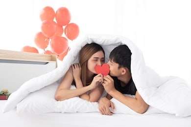 happy young couples hold and kiss a red heart together on a bed under a blanket on morning in the bedroom. young asian couples are enjoying spending time together to celebrate valentine's day.