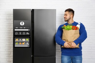 young man with bag of groceries near smart refrigerator indoors