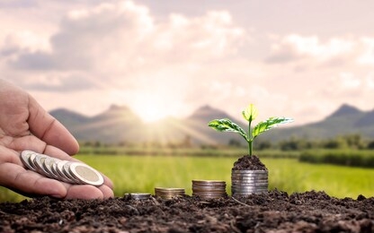 the hand of the farmer holding the coin, there is a small tree planted on a pile of coins or silver with green mountains blurred. the concept of investment for agriculture.