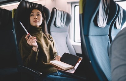pensive asian female author thinking about article creation holding pen and notepad sitting in wagon, young woman student looking away pondering on ideas and plannings spending time on trip in train