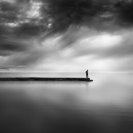 a black and white, fine art image of a man standing on the end of a pier with dramatic sky and still water