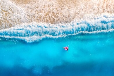 aerial view of a young woman swimming with the donut swim ring in the clear blue sea with waves at sunset in summer. tropical aerial landscape with girl, azure water, sandy beach. top view. travel
