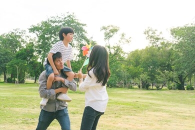 happy asian family playing outdoor activity at park together. son enjoy sitting on father shoulder. mother holding colorful wind turbine. healthy group of people love relationship lifestyle insurance 