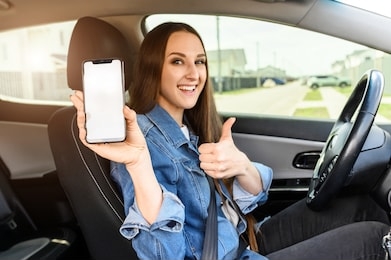 a young happy woman driving a car, she shows empty phone screen and thumb up. useful mobile apps for driver. side view inside car