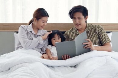 happy asian family with a daughter lying sitting on the bed under the blanket the father is reading a book telling the stories to his daughter.the mother is pat her daughter’s head with love and care