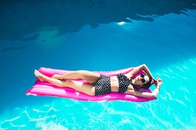 beautiful girl by the pool with long dark hair and make-up resting at a spa resort. in a black retro bathing suit, scarf, hat and glasses, smiles sunbathes on a pink 
inflatable mattress in the motel