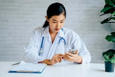 young asian female doctor using a mobile phone at hospital. healthcare worker scrolling and typing text messages at office.