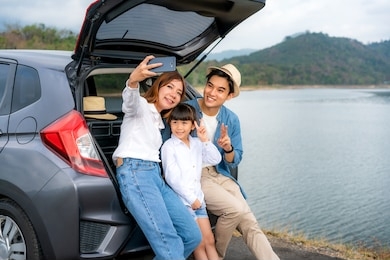 portrait of asian family sitting in car with father, mother and daughter selfie with lake and mountain view by smartphone while vacation together in holiday. happy family time.