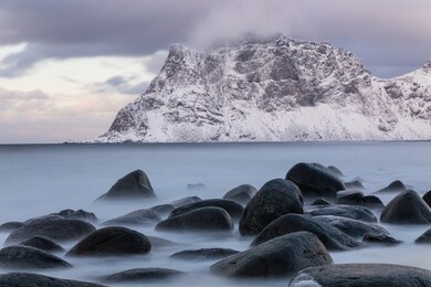 long exposure capture of uttakleiv beach, lofoten, norway. soft sea and rocks. snow covered mountain in background. calm winter scene.