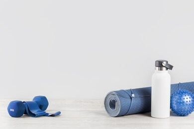 rolled blue yoga mat and white metal water bottle flask grey wooden surface dumbbell rubber resistance band. gender neutral fitness and exercise concept and hydration with copy space. active lifestyle