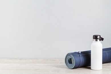 rolled blue yoga mat and white metal water bottle flask on grey wooden surface. gender neutral fitness and exercise concept and hydration with copy space. active lifestyle. workout at home