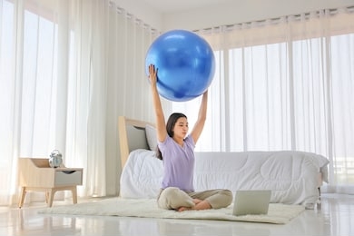 asian lady doing yoga exercise with big ball in her bedroom while watching from online class instructor via laptop as a result of social distancing and quarantine period