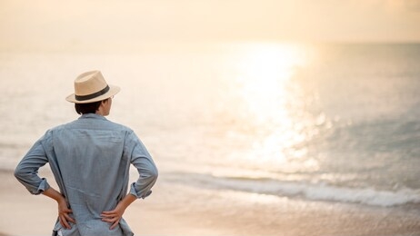 asian man tourist wearing hat standing on tropical island beach enjoy looking at sunset. relaxing holiday or vacation travel in summer season. summertime trip concept