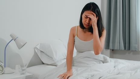 unhealthy sick indian female suffers from insomnia. asian young woman taking painkiller medicine to relieve headache pain and drink glass of water sitting on bed in her bedroom at home in morning.