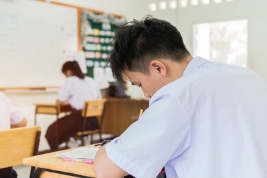 young asian student taking test or final exam, notes information in paper book, concentration of serious in learning at high school classroom with group friends with thai white shirt uniform