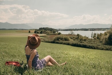 young white woman in a blue dress and brown hat sitting on the grass from back near the lake on a background of mountains in summer.