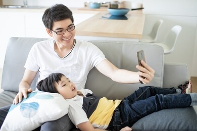 asian happy family father and son using smart phone for video call to mother and sitting in sofa living room