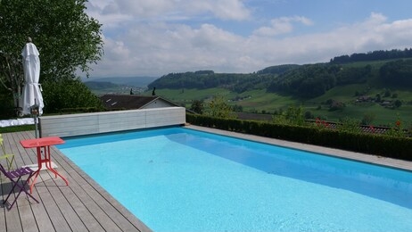 blue pool with landscape in the background, pool with wood floor above. pool with chair and table, sunshine and warm temperature in the summer