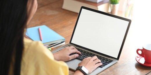 behind shot of beautiful woman working as accountant while sitting and working with white blank screen computer laptop at the wooden working desk that surrounded by coffee cup and stack of books.