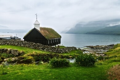 summer view of traditional turf-top church in faroese village. beauty landscape with foggy fjord and high mountains. streymoy island, faroe islands, denmark.