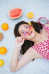 cute beautiful young asian woman wearing fashion eyeglasses and red dress laying on the floor with tropical fruits. summer vacation time.
