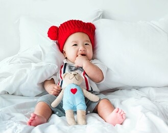 portrait of a newborn asian baby boy, charming child 5 month old wore a suit and a red wool hat sitting in bedroom sucking his finger,fat baby cute and smilingly with a doll,soft selective focus