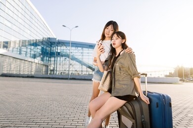two excited pretty young asian brunette girls in casual youth clothes, using smartphone to check their flight or enjoying social networks, waiting for their trip on the airport background