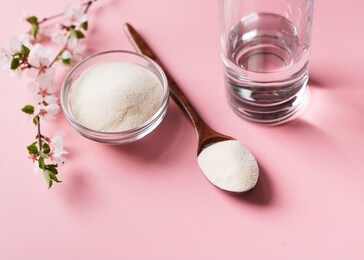 collagen powder in wooden spoon, supplement with galss of water and flowers, healthy and anti age concept  on pink background, top view, copy space