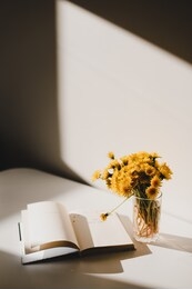 a book and a bunch of yellow dandelions on a white background. a glass vase with dandelions. still life with wild flowers. warm tone. play of light and shadow. good morning. summer floral background