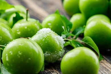 salty green plums with leaves on wooden background, close-up.