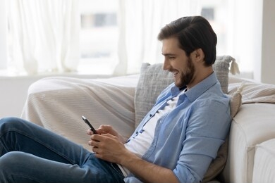 smiling millennial caucasian man sit relax on comfortable couch in living room texting messaging on smartphone, happy young male rest on sofa at home, browse internet, use cellphone gadget