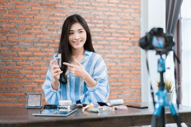asian businesswoman working from home live video interaction with customers using camera device vlogging selling make up products, viral internet social media influencer interacting to live audience