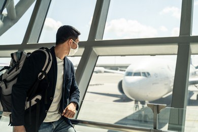 asian traveler business man wearing face mask waiting to board into airplane, standing in departure terminal in airport. male passenger traveling by plane transportation during covid19 virus pandemic.