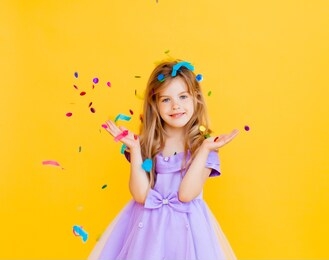 happy little girl with blond hair and in a blue dress catches confetti on a yellow background, holiday concept