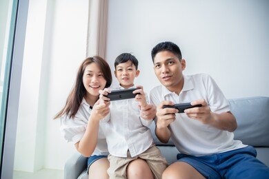 asian family having fun playing computer console games together, father and son have the handset controllers and the mother is cheering the players at home