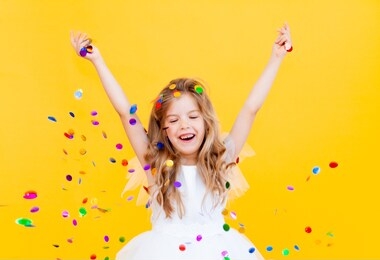 happy little girl with blond hair and in a white dress catches confetti on a yellow background, holiday concept