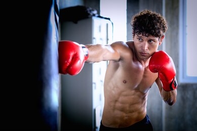 portrait of a confident young athlete caucasian man in boxing gloves, punching a bag in professioal gym. sportsman muay thai boxer work out, training, fighting in gloves.