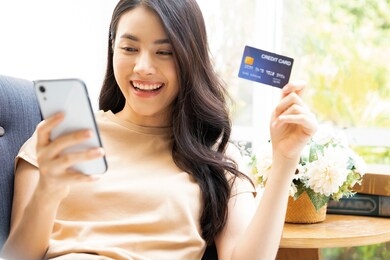 attractive asian woman sitting relaxed on sofa, holding credit card and smartphone probably buying something online.