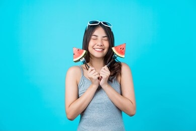 portrait attractive asian young woman holding two piece slide of watermelon and wearing fashion sunglasses on isolated blue color background, copy space and studio, diet and healthy fruit concept