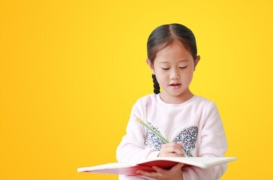 asian little kid girl writes in book or notebook with pencil, isolated on yellow background
